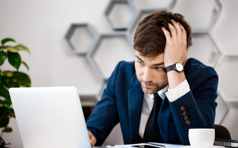 Upset young businessman in suit sitting at laptop at workplace, office background.
